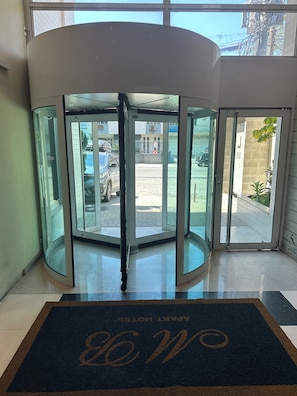 A revolving door with a glass panel, a doormat with a monogram, and a view of a parking area with a building in the background.