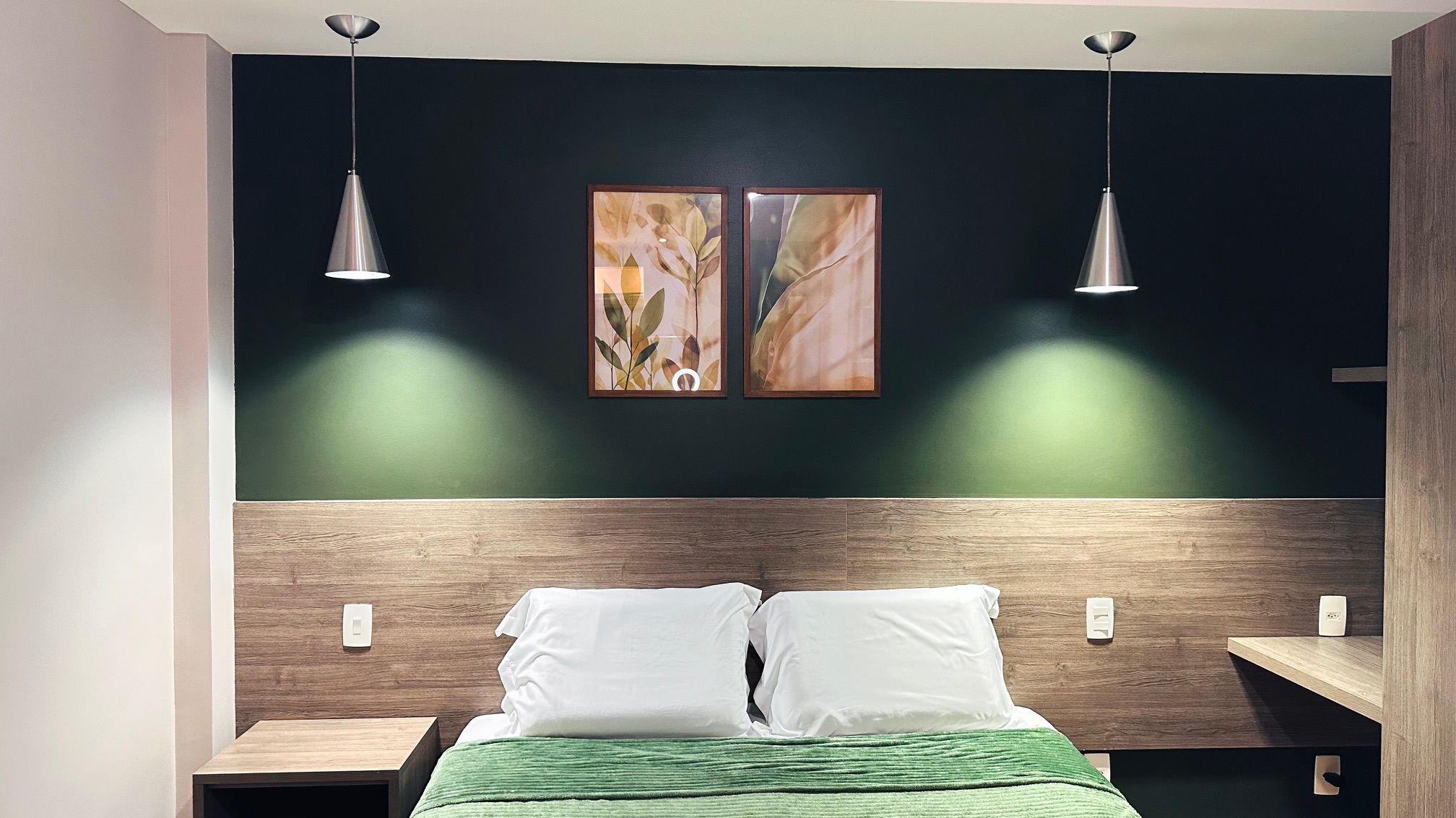 A bedroom with a bed, two bedside tables, a nightstand, a headboard, two pendant lights, and two framed artworks on the wall.