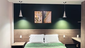 A bedroom with a bed, two bedside tables, a nightstand, a headboard, two pendant lights, and two framed artworks on the wall.