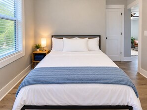 Cama bem arrumada com roupa de cama branca e manta azul, luminária de cabeceira e janela com persianas.