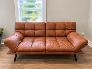 Um sofá de couro marrom com base de madeira e pernas pretas, posicionado em um quarto com uma janela e uma planta verde.