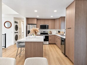 A modern kitchen with wooden cabinets, stainless steel appliances, and a central island with bar stools. - Modern Kent 3BR/2BA Retreat | Kitchen & Laundry (Kent)