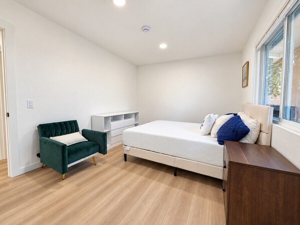 A bedroom with a bed, a green armchair, a white bedside table, and a window with a view.