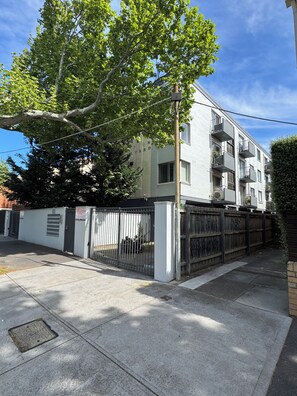 A multi-story residential building with a gated entrance, a tree in front, and a clear sky.