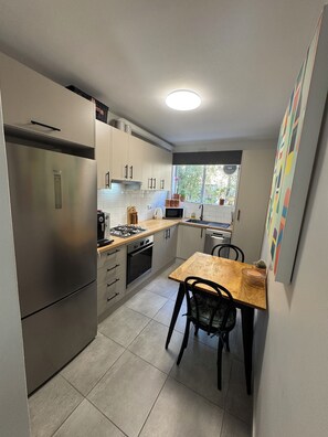 A compact kitchen with a refrigerator, stove, microwave, and sink. There is a dining table with chairs and a window with a view of greenery outside.