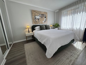A bedroom with a bed, two bedside tables, a lamp, a potted plant, and a painting on the wall.