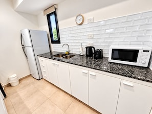 Una cocina moderna con armarios blancos, encimera de granito negro, pileta de acero inoxidable, refrigerador blanco, microondas, cafetera y un reloj en la pared.