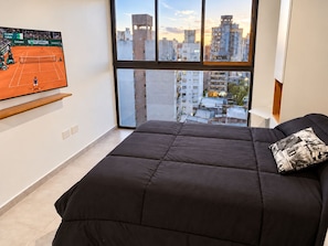 Un dormitorio con un ventanal grande, una cama con cubrecama negro y un televisor de pantalla plana mostrando un partido de tenis.