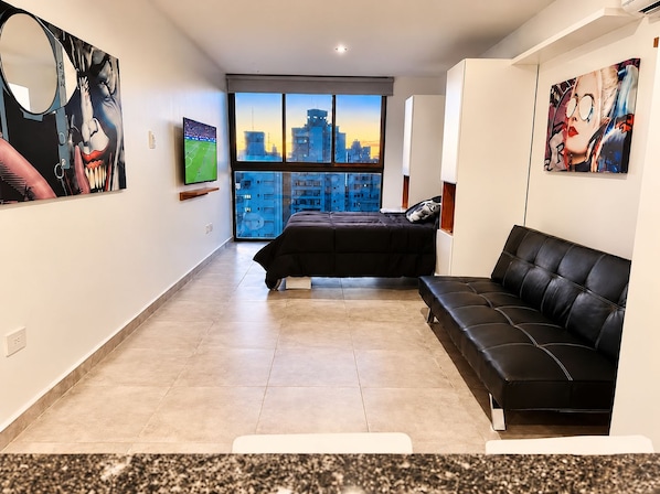 Un dormitorio moderno con cama, sofá de cuero negro, ventana grande con vista a la ciudad y obras de arte en las paredes.