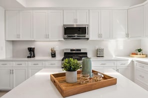 Une cuisine moderne avec des meubles blancs, un four à micro-ondes en acier inoxydable, une machine à café et un plateau en bois sur lequel sont posés une plante et des verres.