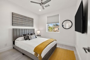 Une chambre à coucher comprenant un lit, un ventilateur de plafond, une fenêtre avec des stores, un téléviseur fixé au mur et une table de chevet avec une lampe.