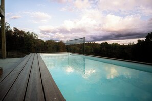 Ein Schwimmbecken mit Holzterrasse und Metallzaun, umgeben von Bäumen und einem bewölkten Himmel.