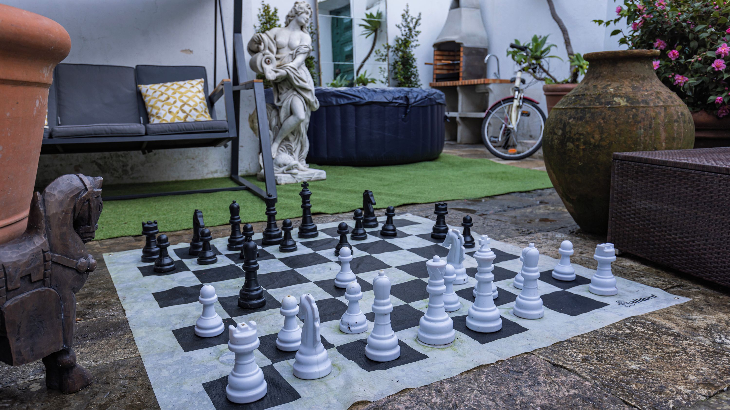Un jeu d’échecs installé à l’extérieur, avec une statue et une chaise en osier en arrière-plan.