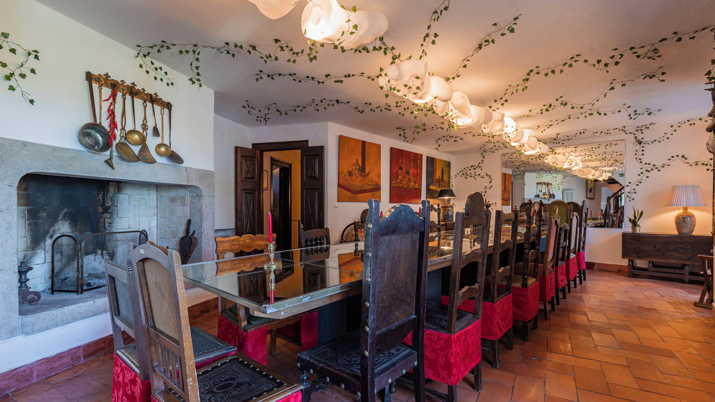 Un espace repas avec une longue table en bois, des chaises recouvertes de tissu rouge, une cheminée et des luminaires suspendus.