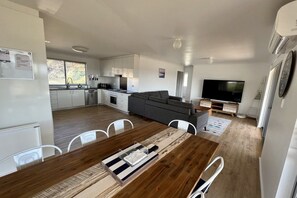 Une cuisine moderne avec des meubles blancs, un coin repas avec une table et des chaises en bois, un canapé gris et un téléviseur à écran plat.
