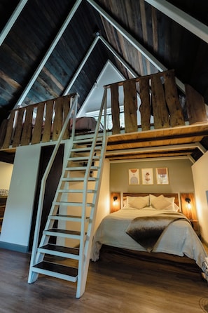 A loft bedroom with a wooden staircase, a bed with pillows, and wall-mounted lighting.
