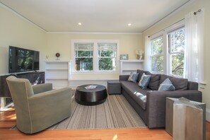 Un salon moderne avec un canapé d’angle, un téléviseur à écran plat et une grande fenêtre équipée de stores.