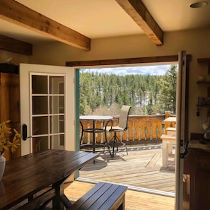 Eine Holzdielen-Terrasse mit Tisch und Stühlen, eine Tür, die zu einem Raum führt, und Blick auf einen Wald.