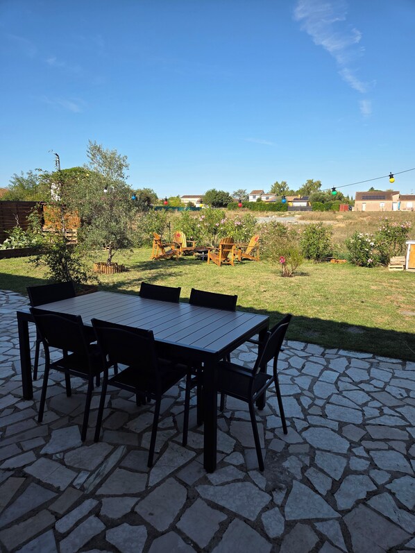 Un patio al aire libre con una mesa y sillas, rodeado de un jardín y vistas a casas a lo lejos.