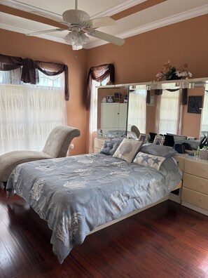 Un dormitorio con cama, tocador con espejo, ventilador de techo y ventana con cortinas.