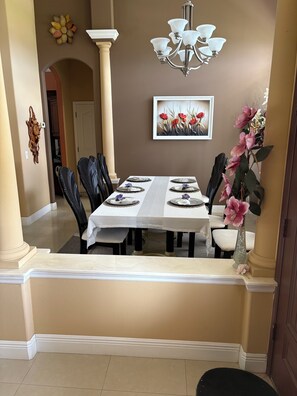 Un comedor con una mesa dispuesta para cuatro personas, flanqueada por sillas, una araña y un cuadro floral enmarcado.