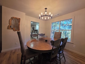 Comedor con mesa y sillas de madera, un candelabro, una ventana con vistas a árboles y una cocina al fondo.