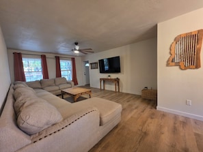Een woonkamer met een hoekbank, een salontafel, een aan de muur bevestigde televisie en een plafondventilator.