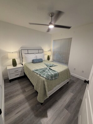 Un dormitorio con cama, mesitas de noche, ventilador de techo y ventana con persianas.