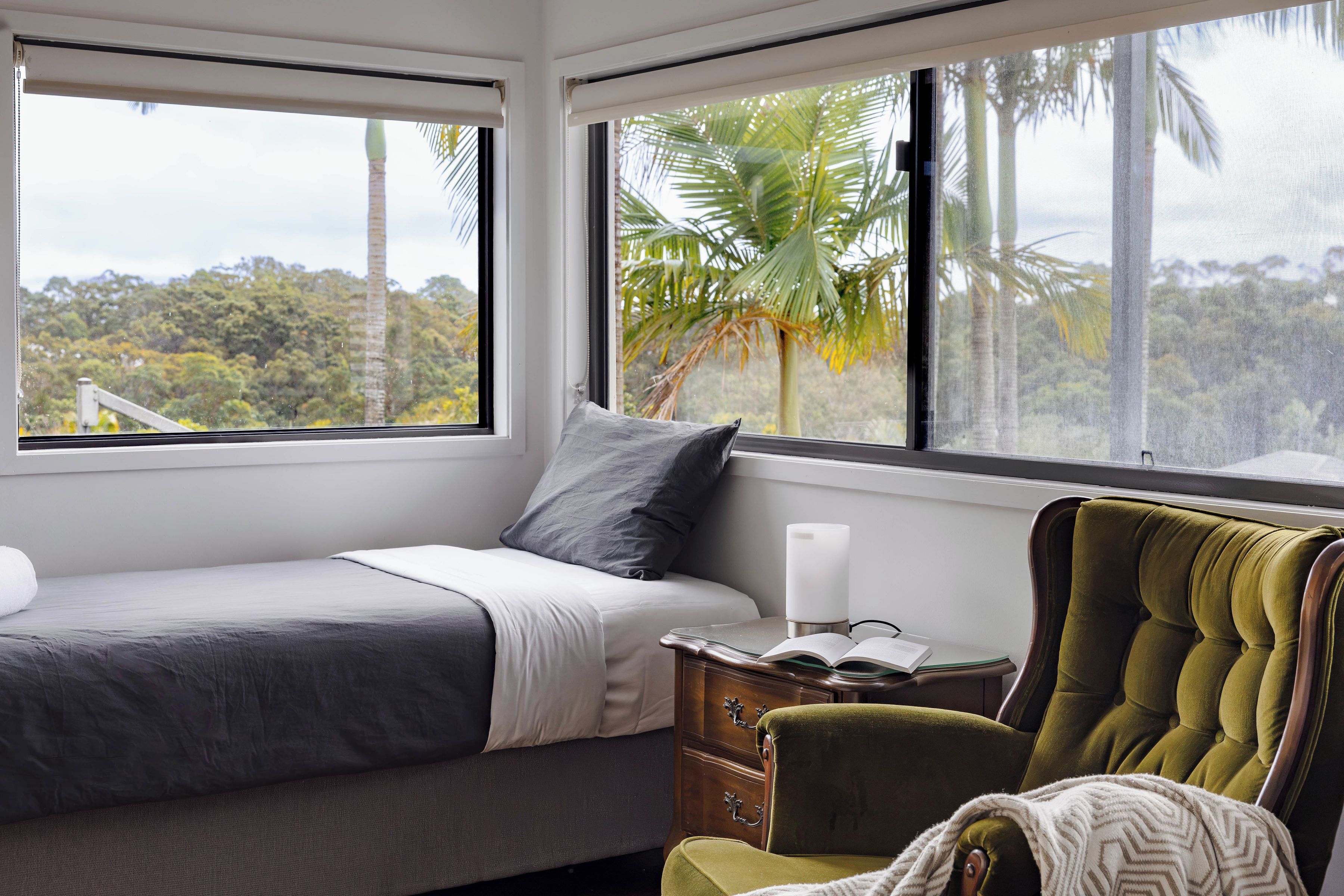 Quarto com cama, cadeira, mesa de cabeceira e janela com vista para palmeiras e uma floresta.
