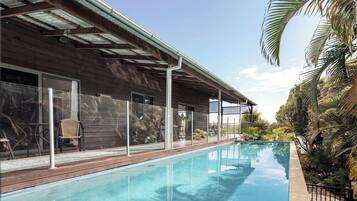 Piscina com deck de madeira, guarda-corpos de vidro e vista para um edifício com telhado metálico.