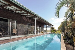Piscina com deck de madeira, guarda-corpos de vidro e vista para um edifício com telhado metálico.