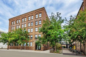 A multi-story brick building with a row of windows on the ground floor, a covered entrance, and a sidewalk with trees and outdoor seating. - Modern Studio in Downtown DSM + Free Parking + Gym + Workspace (Des Moines)