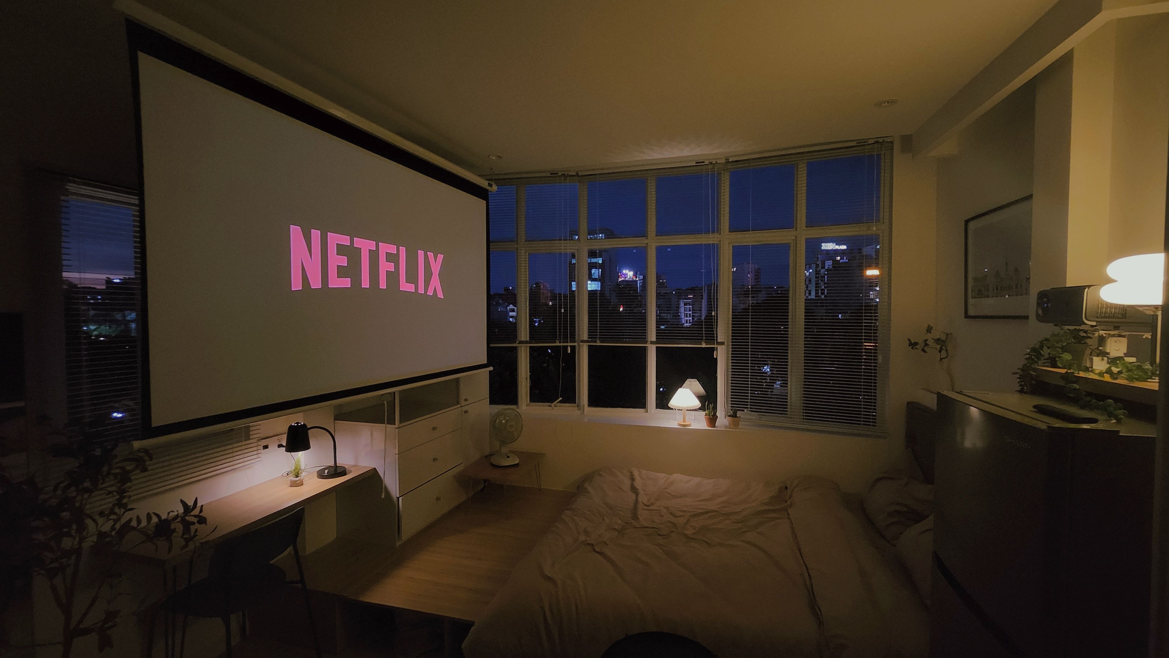 A bedroom with a large window, a bed, a desk with a laptop, and a wall-mounted projector displaying 'NETFLIX'.