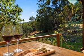 Een houten tafel met een glas rode wijn en een bord eten, met uitzicht op een natuurlandschap met bomen en een heldere lucht.