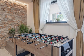 Una mesa de fútbolín en una habitación con pared de piedra, una ventana con cortinas y una planta.