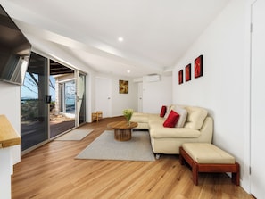 Un salon moderne avec un plancher en bois, un canapé d’angle beige, une table basse et une porte-fenêtre coulissante donnant sur un espace extérieur.