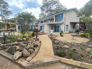 Une maison moderne avec une façade en pierre, une allée pavée et un jardin agrémenté de rochers et de plantes.
