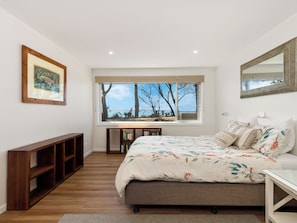 Une chambre à coucher avec un grand lit, un bureau, une bibliothèque et une fenêtre donnant sur les arbres et le ciel.