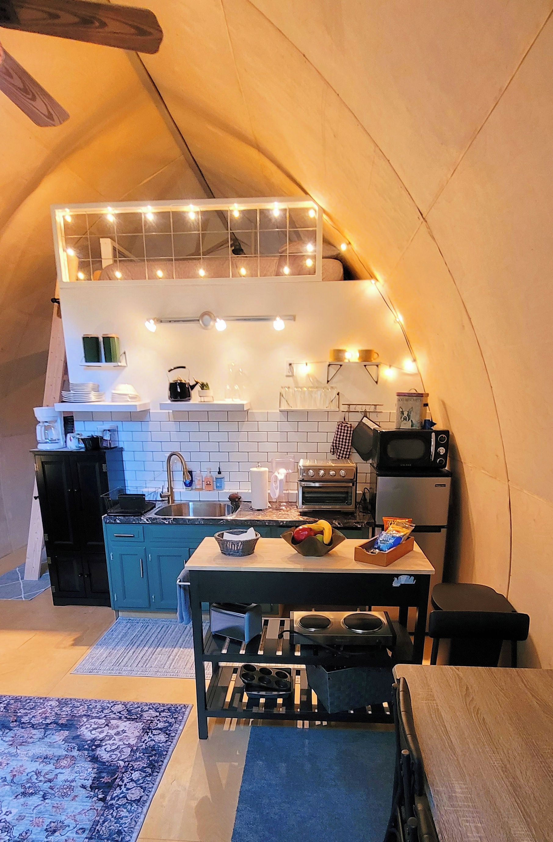 A compact kitchen with a sink, stove, and microwave, under a tent with string lights.