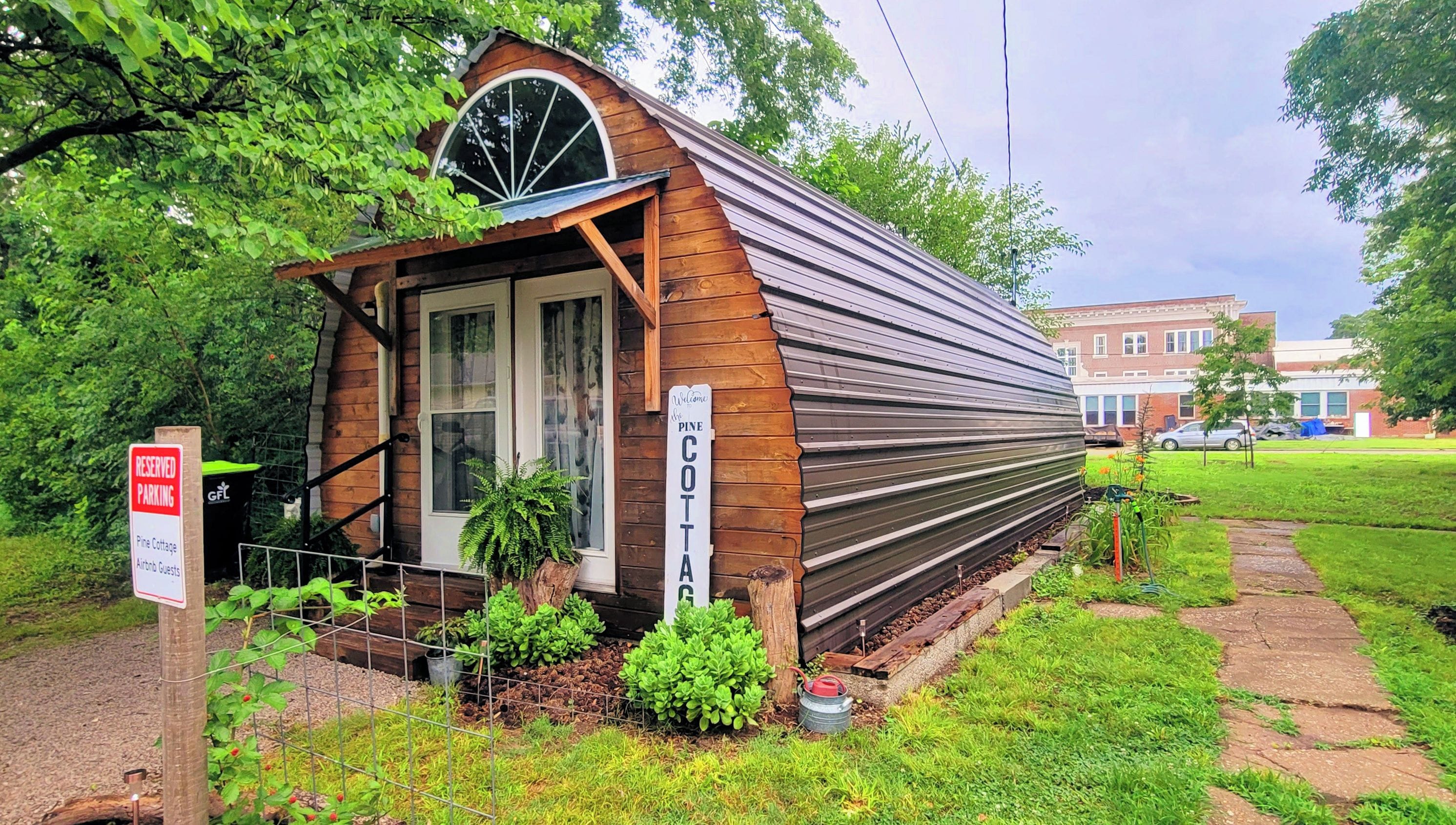 A small, rustic cabin with a welcoming sign, surrounded by lush greenery and a paved path.