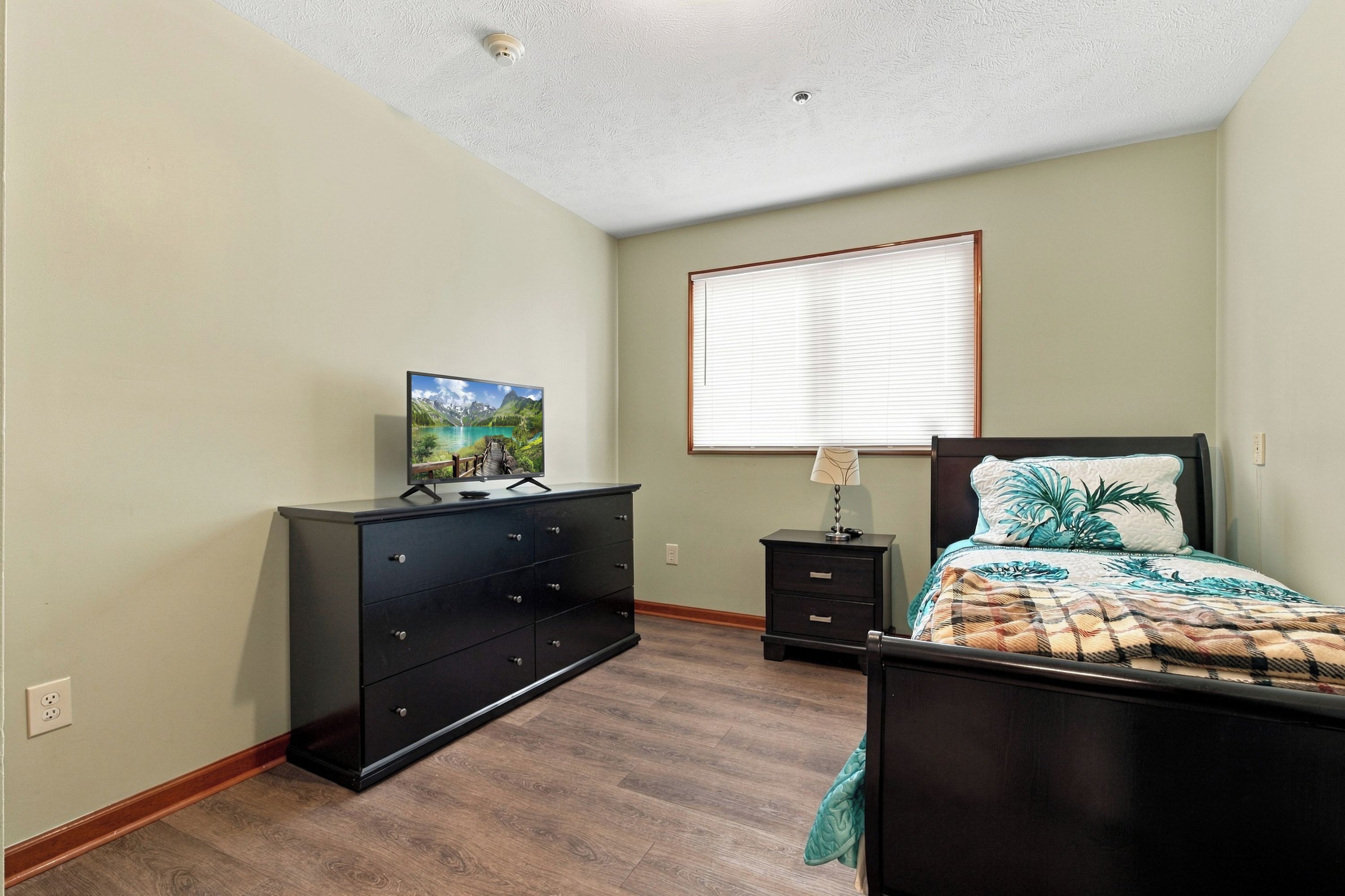 Une chambre à coucher avec un lit, une commode, une table de chevet, une lampe et un téléviseur posé sur la commode.