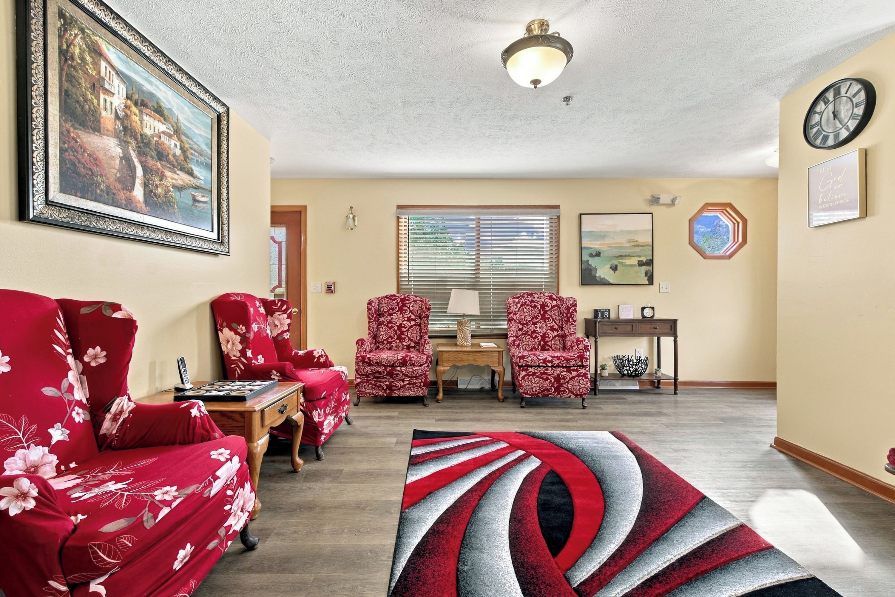 Un salon avec un canapé à motifs floraux rouges, deux fauteuils à motifs, une table basse, un tapis, un tableau, une horloge et une fenêtre avec des stores.