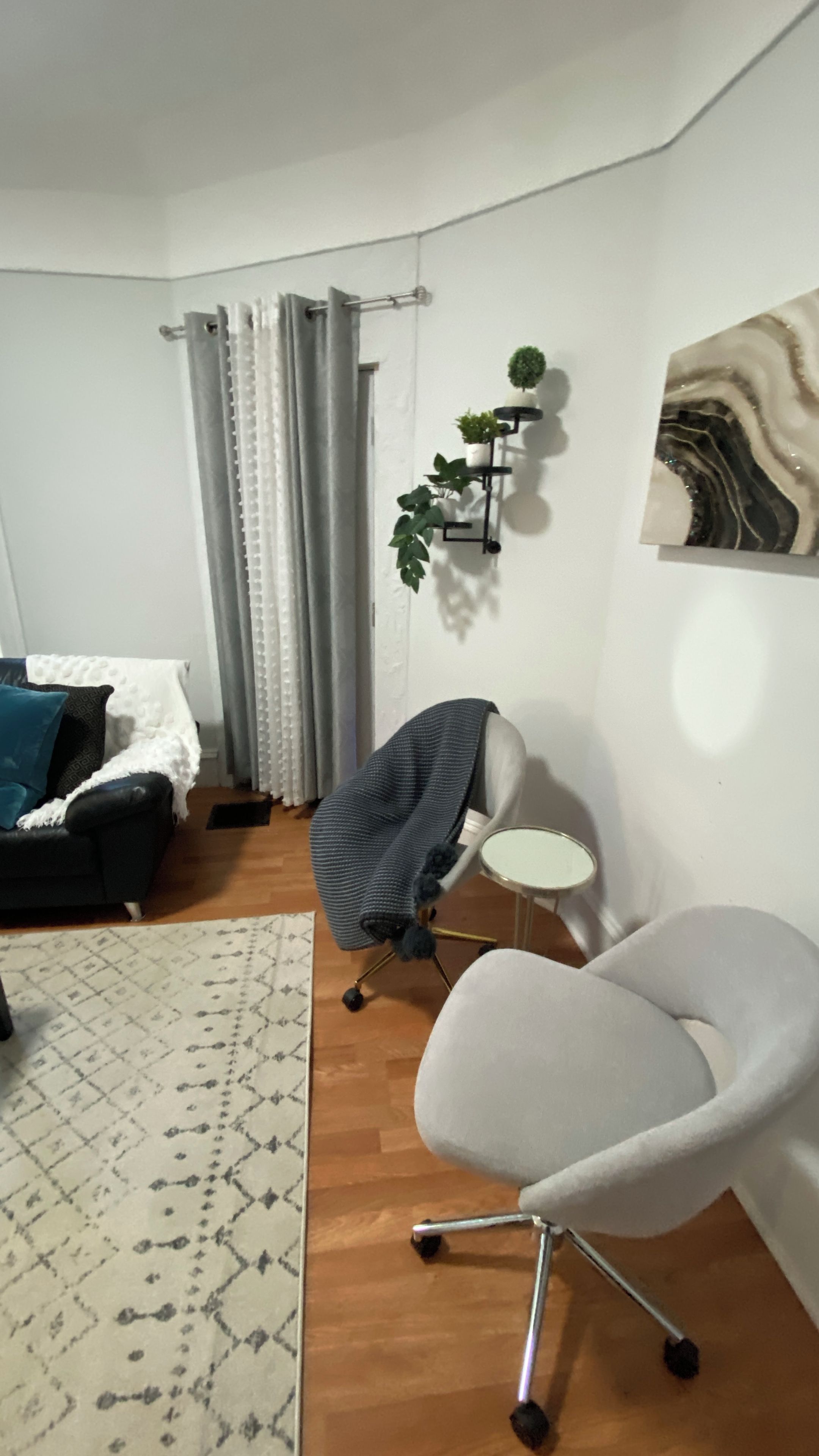 Una sala de estar con un sofá, un sillón gris, un tapete estampado, una estantería para plantas montada en la pared y un cuadro.