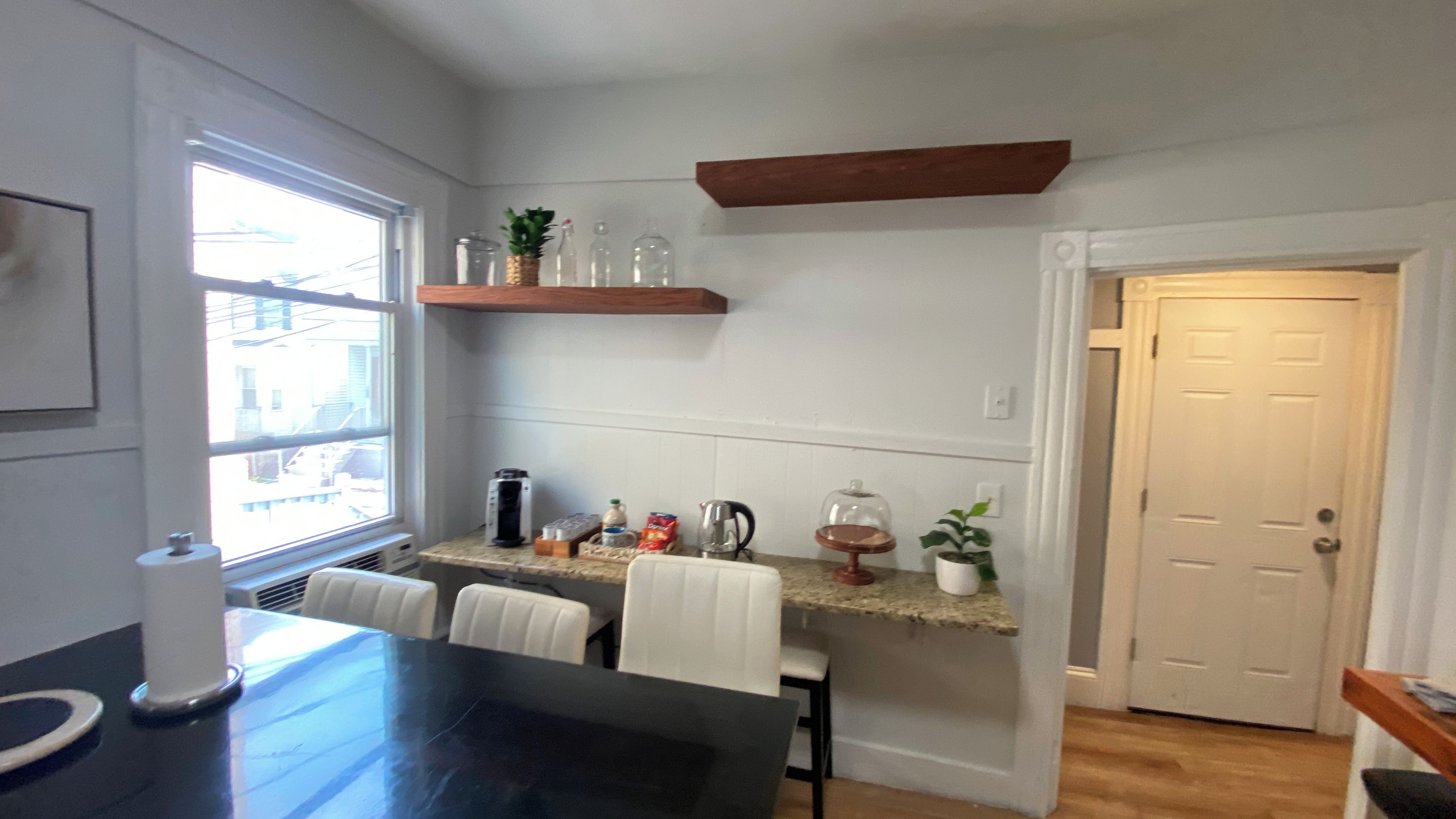 Una cocina con una mesa de comedor, sillas, una cafetera y una estantería con frascos y una planta.