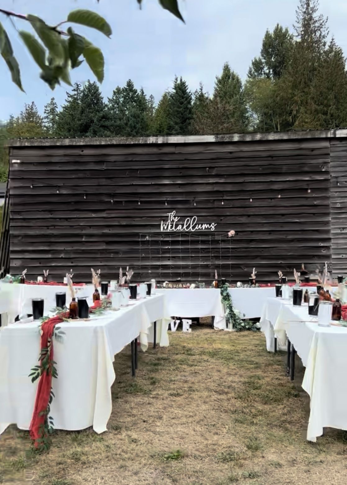 An outdoor event space with tables set up for a reception, featuring a rustic wooden backdrop with the text 'The Willums.'