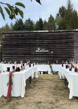 Un espace extérieur aménagé pour les événements, avec des tables disposées pour un cocktail, et en arrière-plan, un décor rustique en bois sur lequel on peut lire le texte : « The Willums ».