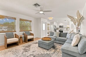 Ein modernes Wohnzimmer mit einem grauen Ecksofa, zwei Holzstühlen, einem runden Hocker und einem Teppich mit Muster.