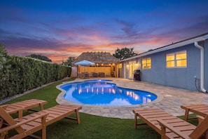 Ein Swimmingpool mit einer strohgedeckten Struktur, eine Terrasse mit Sitzgelegenheiten und Blick auf einen Sonnenuntergangshimmel.