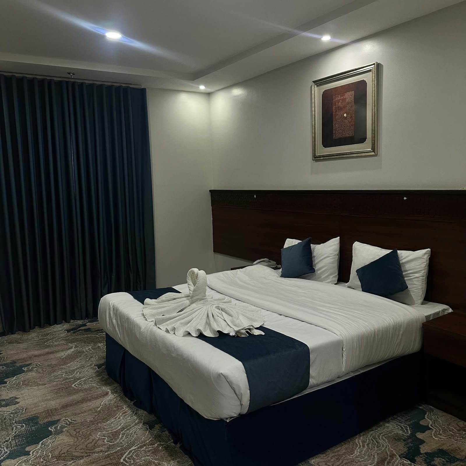 Een hotelkamer met een groot bed, donkere gordijnen, een ingelijst schilderij aan de muur en een kamerbreed tapijt met dessin.