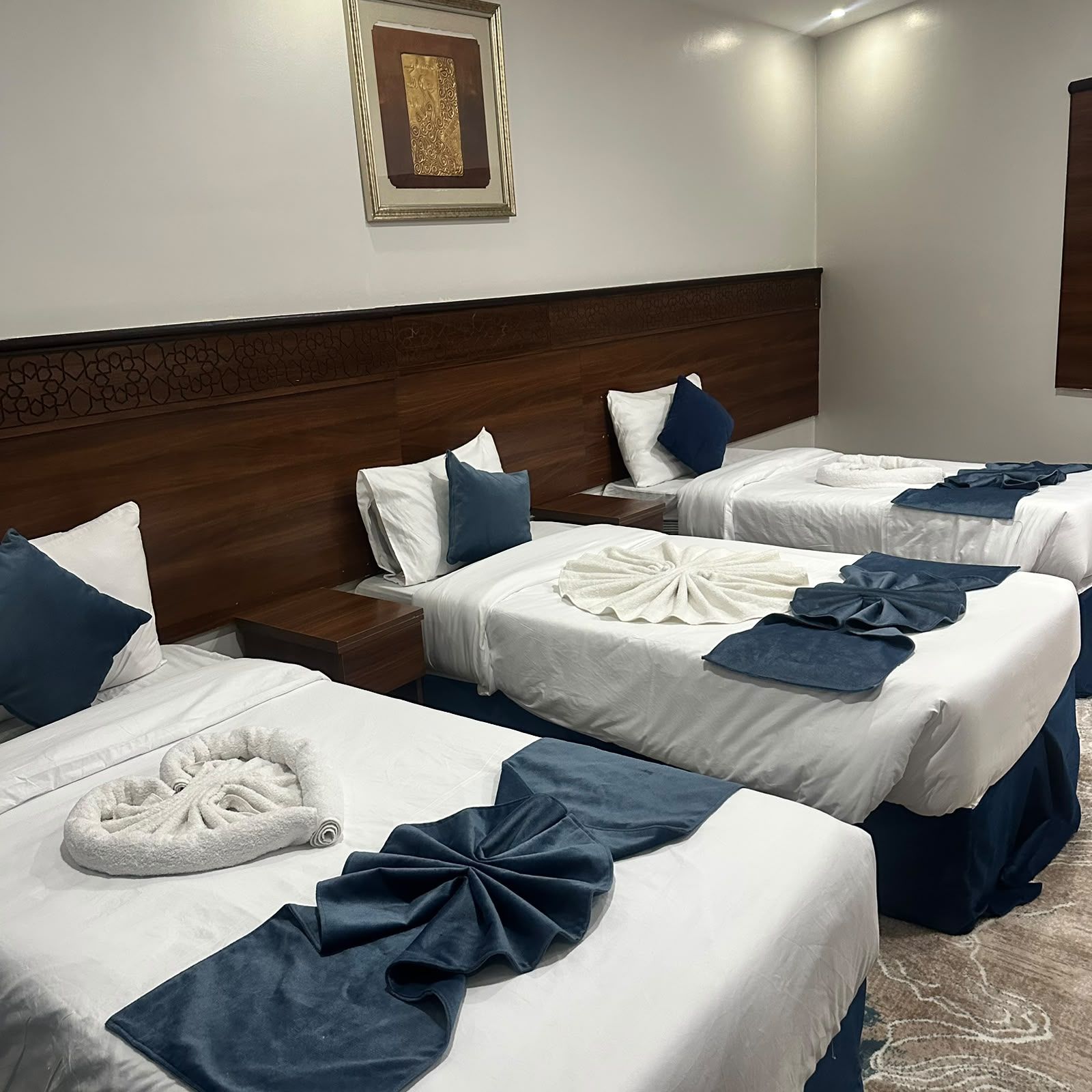 Une chambre d’hôtel avec trois lits, chacun agrémenté de draps blancs et de touches de bleu, d’une tête de lit en bois et d’une œuvre d’art encadrée au mur.